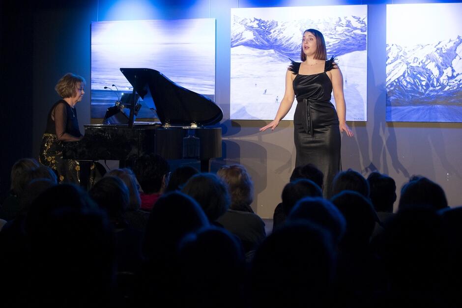 kobieta w czarnej sukni śpiewa, obok mężczyzna przy fortepianie