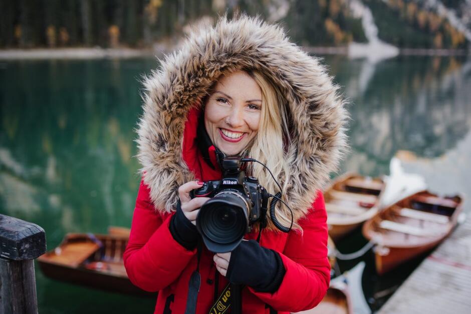 Dziewczyna w czerwonej kurtce i kapturze uśmiecha się do zdjęcia, trzyma w dłoniach aparat fotograficzny.