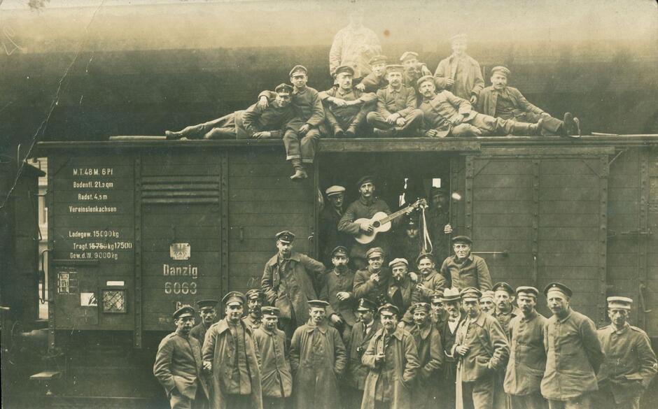 Kilkudziesięciu mężczyzn w mundurach stoi przed wagonem, kilku leży na dachu wagonu