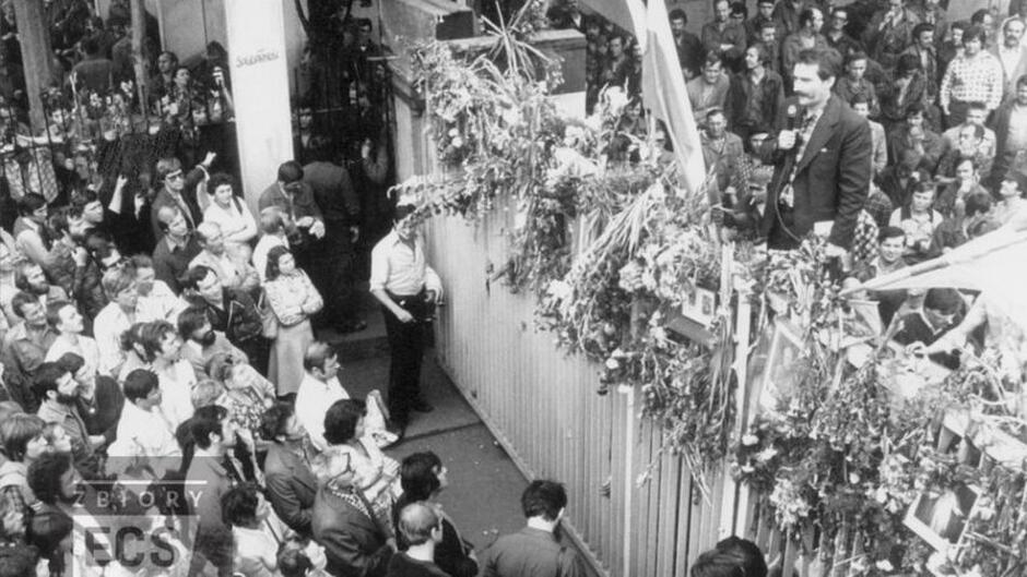 historyczna brama prze stocznią, po jednej tłum mieszkańców, po drugiej strajkujący, na płocie mężczyzna z wąsem