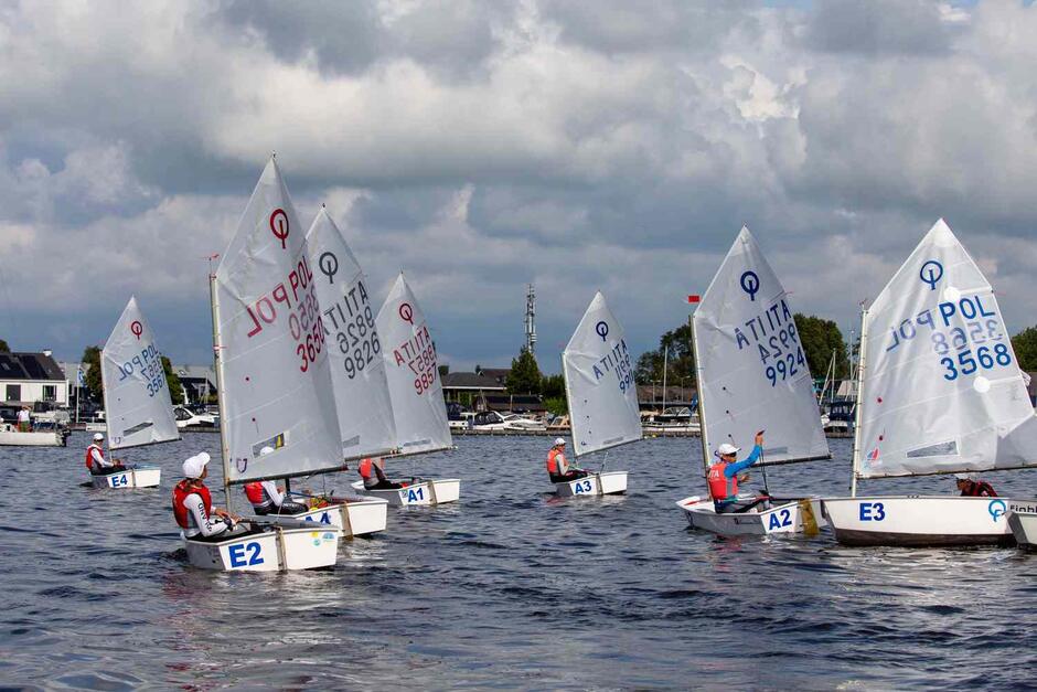 Siedem małych łódek z żaglami na wodzie, w każdej siedzi jeden młody żeglarz/żeglarka w kapoku. Płyną obok siebie 