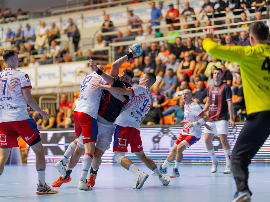 Zawodnik w ciemnej koszulce z piłką w ręku u góry, próbuje przedrzeć się przez dwóch zawodników w jasnych koszulkach. Po prawej bramkarz w żółtej bluzie