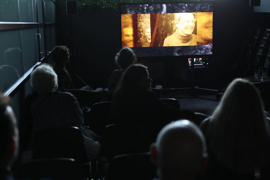 na zdjęciu przyciemniona sala, na krzesłach siedzi kilka osób oglądają film wyświetlany przed nimi na dużym ekranie