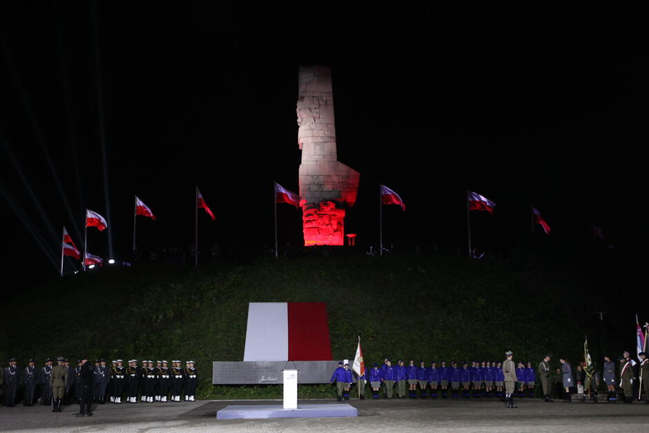 Ciemnawo, wzgórze z podświetlonym na biało i czerwono pomnikiem. Pod nim w rzędzie stoją żołnierze 