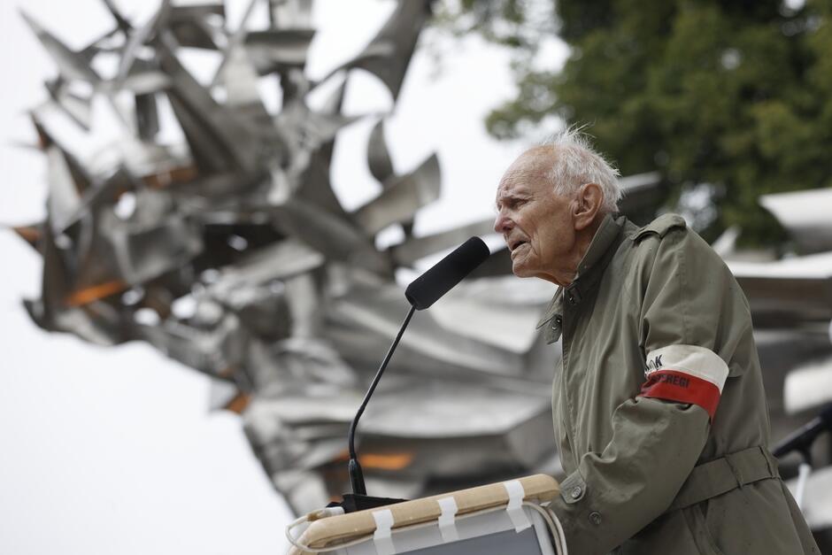 starszy mężczyzna z biło-czerwoną opaską przemawia na deszczu pod metalowym pomnikiem