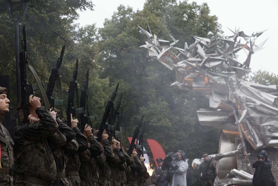 kompania wojska w deszczu na tle pomnika oddaje salwę honorową