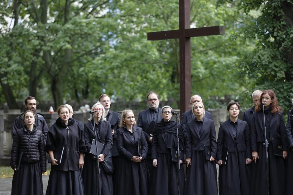 Chórzystki i chórzyści w czarnych strojach stoją pod krzyżem i śpiewają