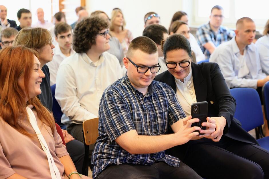 Chłopiec z autyzmem pokazuje telefon komórkowy kobiecie w okularach. Obok po lewej inna uśmiechnięta kobieta, w tle inne dzieci
