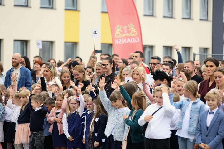 Dzieci i dorośli odświętnie ubrani przed szkołą, nad nimi czerwony sztandar z logo Miasta Gdańska
