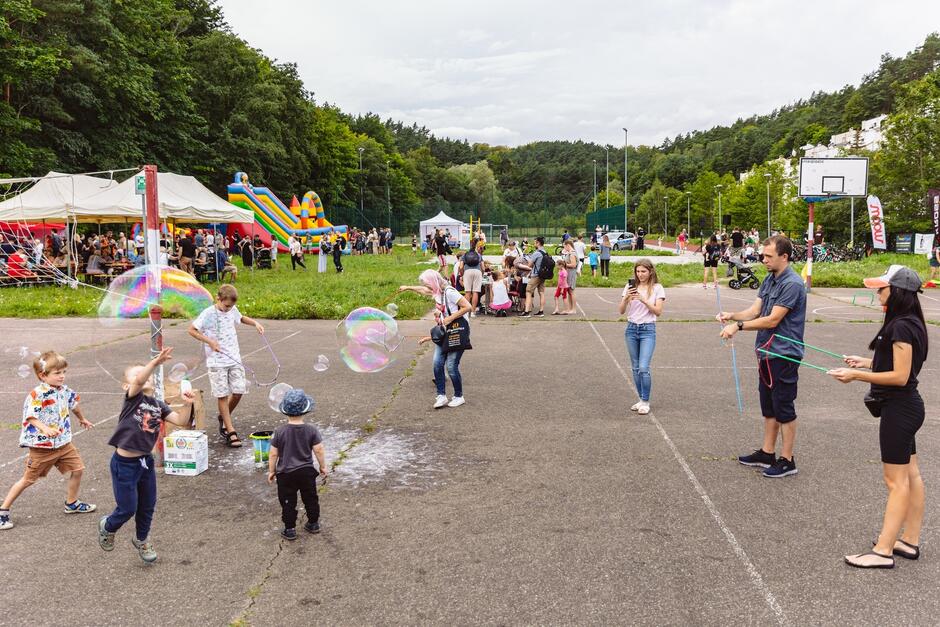 na zdjęciu grupa kilkorga dzieci, bawią się z dużymi bańkami mydlanymi na boisku szkolnym
