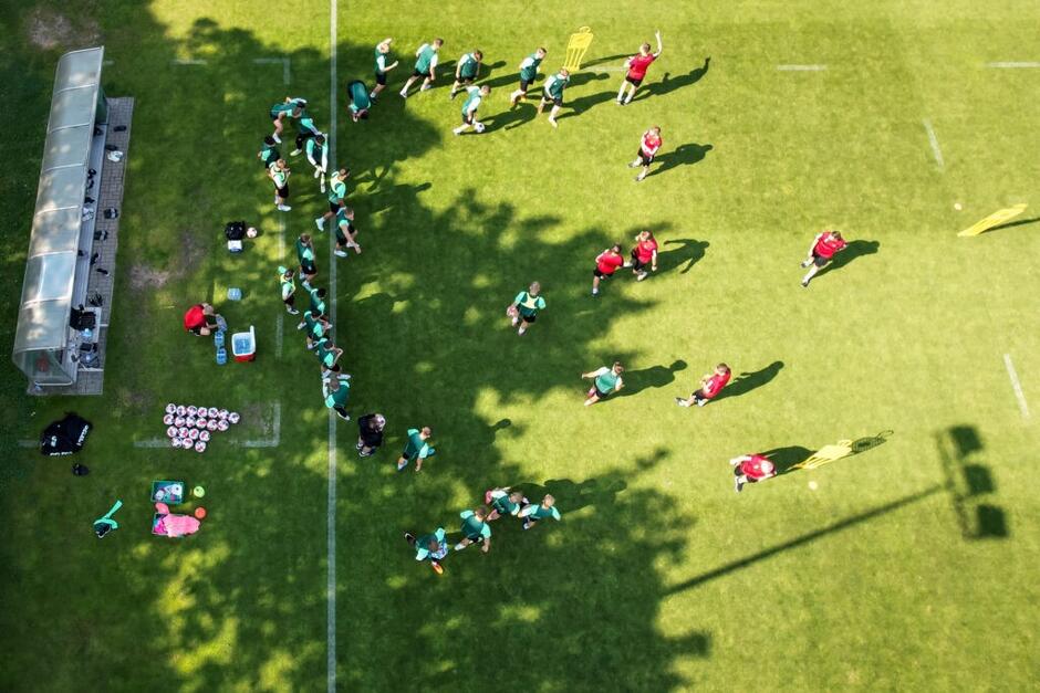 Wykonane z wysokości kilkunastu metrów, przy pomocy drona, zdjęcie trawiastego boiska, po którym biegają piłkarze 