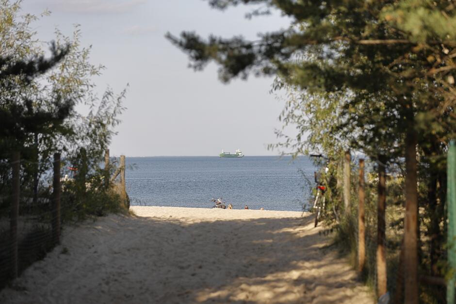 na zdjęciu wejście na plaże, widać piasek, po jego obu stronach drzewa, a w tle wodę morską