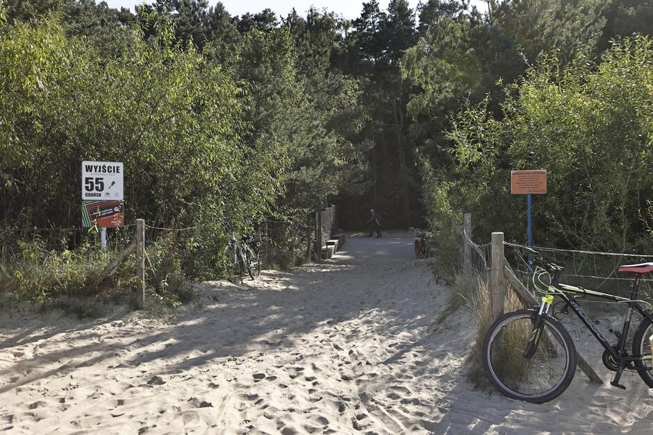 na zdjęciu wejście na plażę, widać dużo piasku, a po obu stronach wejścia zielone drzewa, po prawej stoi rower