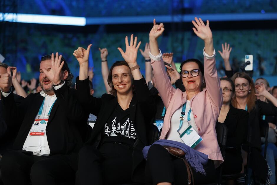 Fragment widowni, osoby siedzące w rzędach. W pierwszym rządzie - trzy osoby z radośnie uniesionymi w górę rękami. Od prawej: kobieta, kobieta, mężczyzna 