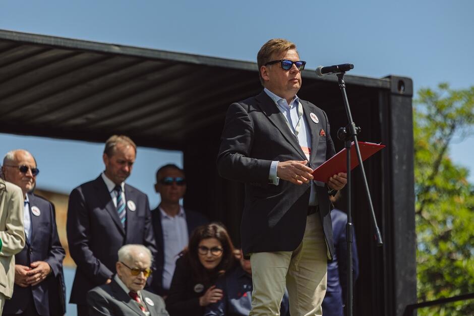 mężczyzna w średnim wieku, w marynarce przemawia do mikrofonu w tle siedzą różne osoby