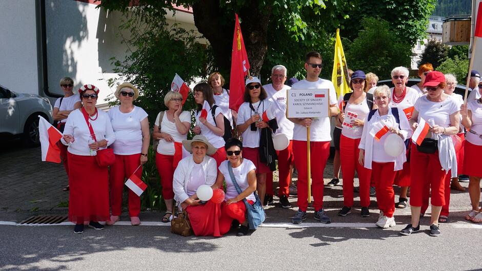 osoby ubrane na biało czerwono pozują do zdjęcia z tabliczką z nazwą chóru i flagą Polski