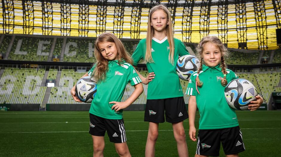 Trzy dziewczyny w różnym wzroście w biało-zielonych strojach stoi na murawie, pod ręką każda trzyma piłkę do piłki nożnej. W tle trybuny z napisem LECHIA