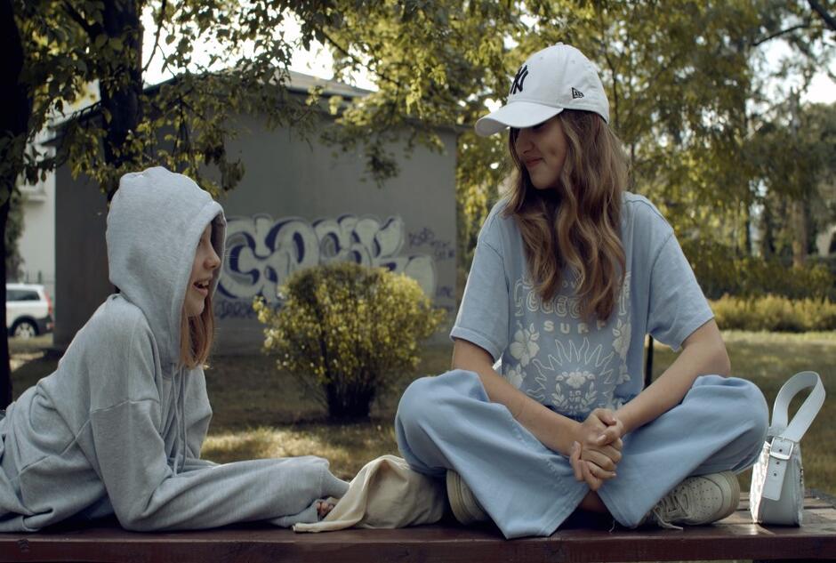 Dwie dziewczynki na ławce, jedna jest ubrana w bluzę z kapturem, druga ma koszulkę i czapkę z daszkiem - rozmawiają