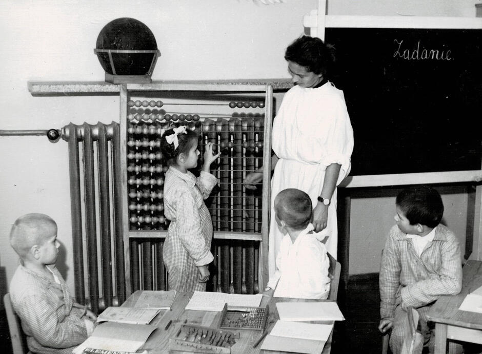 Zdjęcie czarno-białe, dziecko stojące przed dużym liczydłem i tablicą szkolną, obok nauczycielka i troje uczniów siedzących w ławkach