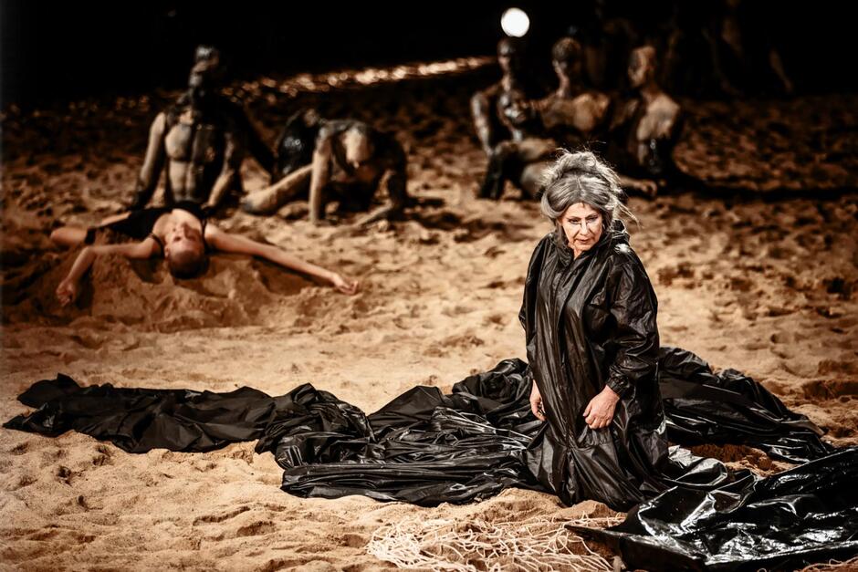 kobieta w siwej peruce i ogromnej czarnej sukni siedzi na scenie wysypanej piaskiem