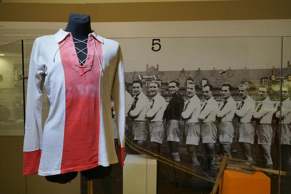 Zdjęcie przedstawia koszulkę piłkarską KS Gedania z długimi rękawami. Środek jest czerwony, boki i rękawy są białe. W tle czarno-białe zdjęcie przedstawiające sportowców z dwudziestolecia międzywojennego ustawionych w szeregu spoglądających na widza.