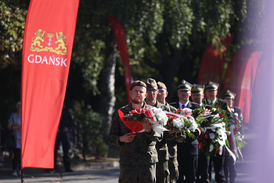 Grupa żołnierzy niosących wieńce na tle flagi Gdańska