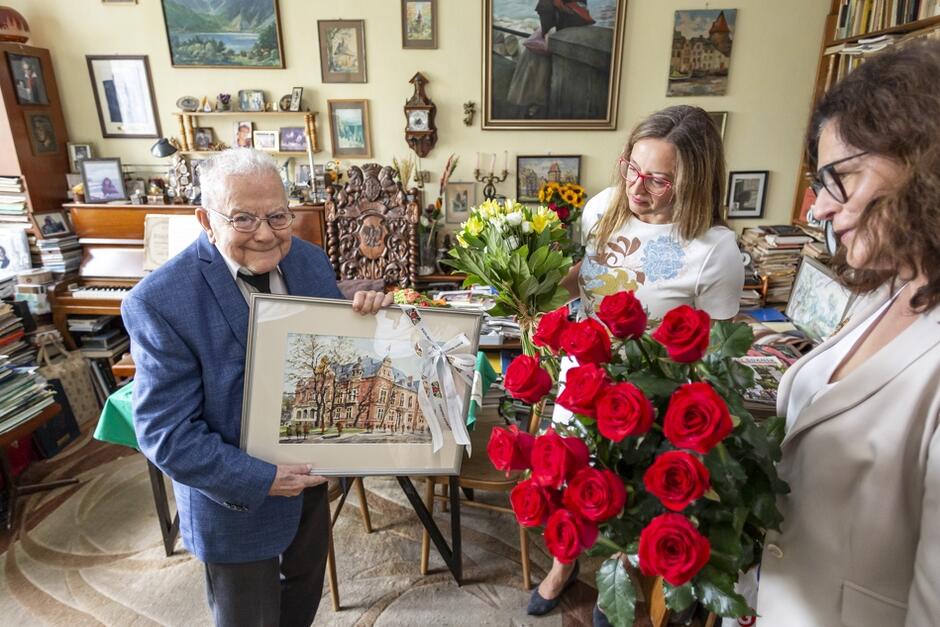 starszy mężczyzna odbiera obraz, obok dwie kobiety, jedna z bukietem czerwonych róż