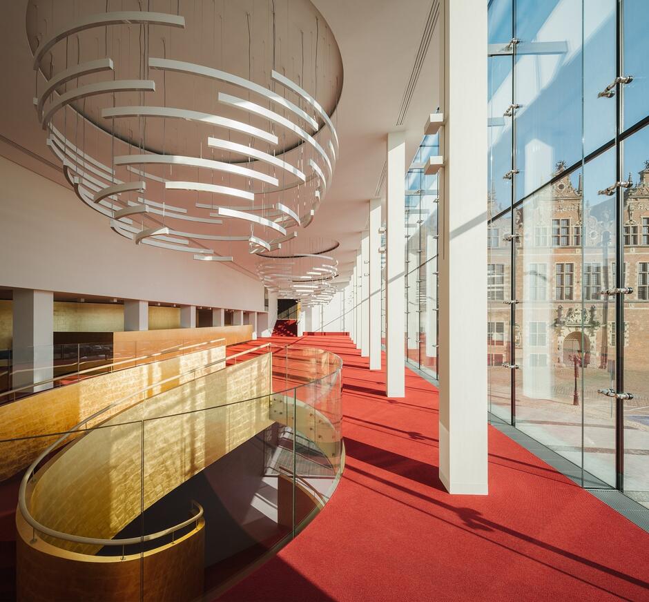 wnętrze z innego kąta, czerwona wykładzina i duży nowoczesny żyrandol, po prawej szklana fasada, po lewej złota balustrada schodów prowadzących w dół