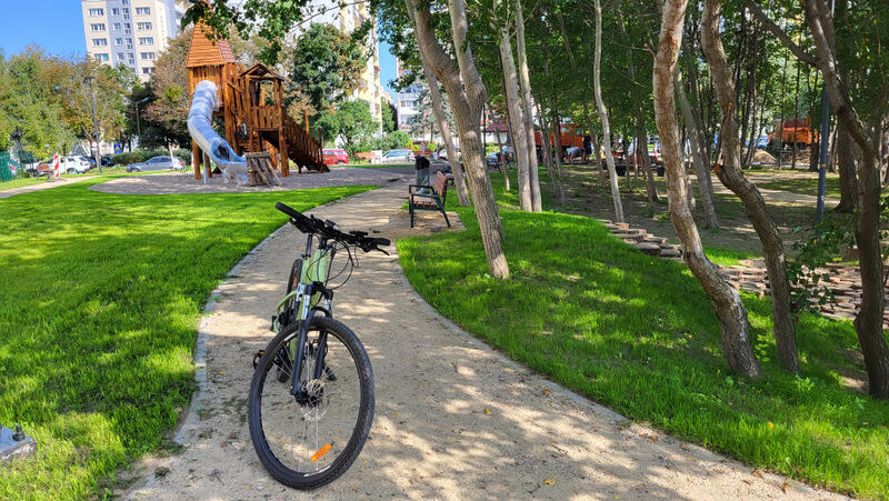 fragment parku: drzewa, ścieżka z rowerem, trawnik, zjeżdżalnia 