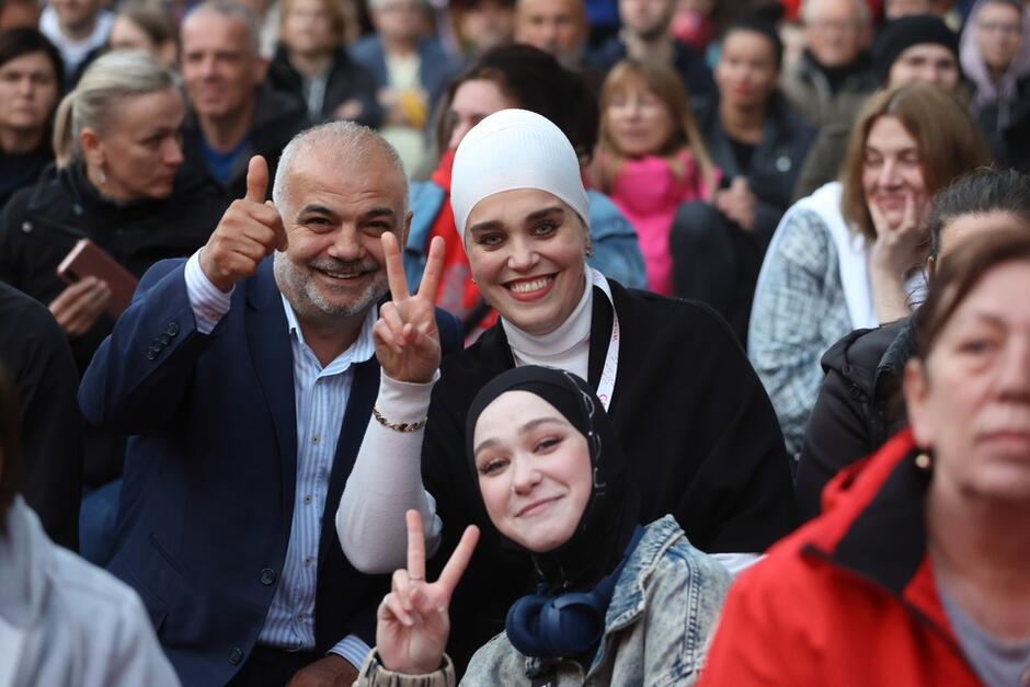 Troje osób tłumie: uśmiechają się, kobiety mają na głowach chusty, wznoszą dwa palce na znak victorii