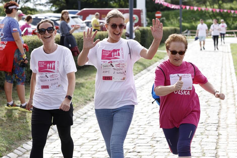 Trzy kobiety biegną - mają w rękach kije do nordic walking i różowe i białe koszulki, są roześmiane