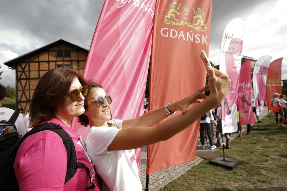 Dwie kobiety - w różowej i białej koszulce, stoją na tle flag Gdańska i fundacji. Robią sobie selfie