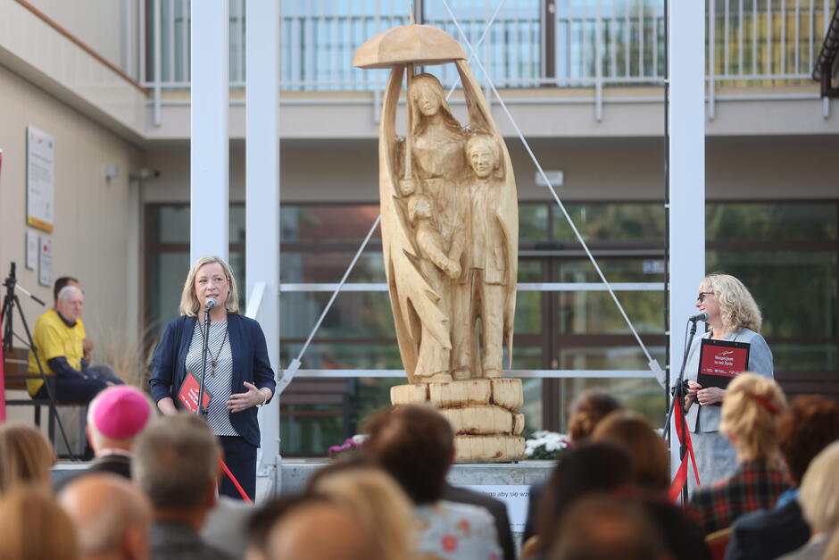 Dwie kobiety stoją przy mikrofonach, za nim potężna rzeźba w drewnie. To postać anioła