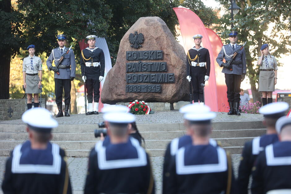 oddział marynarki wojennej, a w tle kamień stojący na skraju parku z napisem Armia Krajowa , obok warty żołnierzy i harcerzy