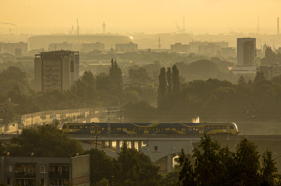 poranne zdjęcie: budynki mieszkalne, tory po których jedzie pociąg, drzewa, w dalekiej perspektywie stadion Polsat Plus Arena Gdańsk