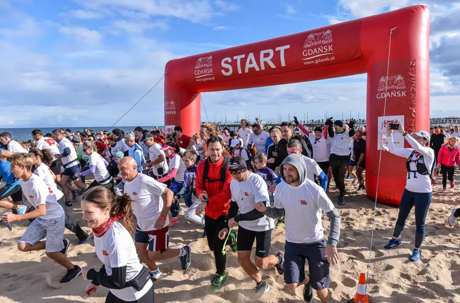 na zdjęciu biegacze, biegną po plaży, w sportowych strojach, widać za nimi wielką czerwoną dmuchaną bramę z napisem start