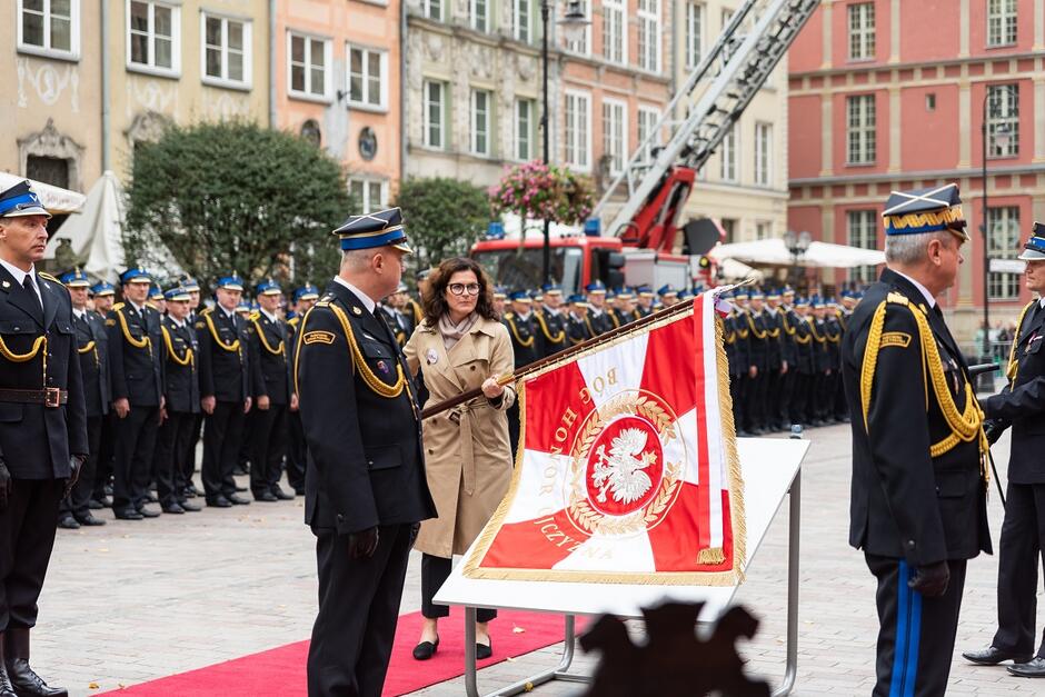 sztandar z napisem Bóg honor ojczyzna uniesiony przez prezydent w kierunku stojącego przed nią strażaka, w szeregu stoją strażacy