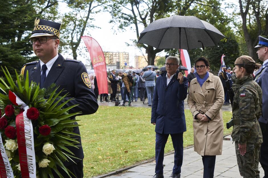 Uroczystość na cmentarzu z udziałem wielu osób. Wieniec znajduje się po lewej stronie zdjęcia, jest niesiony przez funkcjonariusza Straży Miejskiej. Za nim idą dwie osoby - prezydent Gdańsk i radny, który niesie parasol