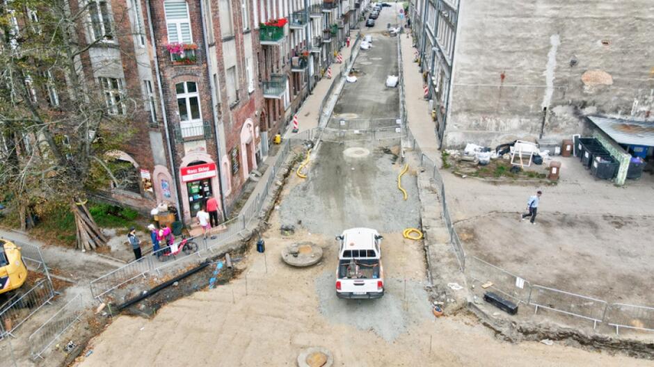 zdjęcie z drona, widać rozkopaną ulicę, na której prowadzone są prace, stoi tam samochód osobowy, widać kilka barierek i fragmenty kamienic