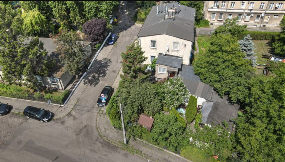 na zdjęciu z drona zabudowania niewysokie, to niezbyt ładne szare budynki, widać fragment ulicy z zakrętem i kilka zaparkowanych samochodów