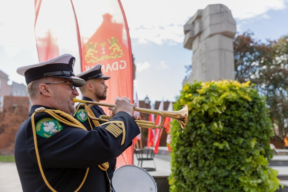 Dwaj żołnierze w mundurach Marynarki Wojennej na tle pomnika i baneru z napisem Gdańsk. Żołnierz znajdujący się na pierwszym planie dmie w trąbkę