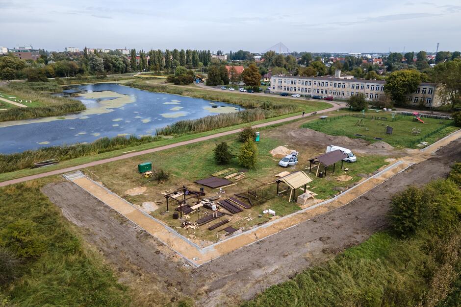 na zdjęciu z drona widać plac budowy pola biwakowego, w tle płynąca rzeka Motława, po prawej w oddali widać budynek szkoły, dwukondygnacyjny szary
