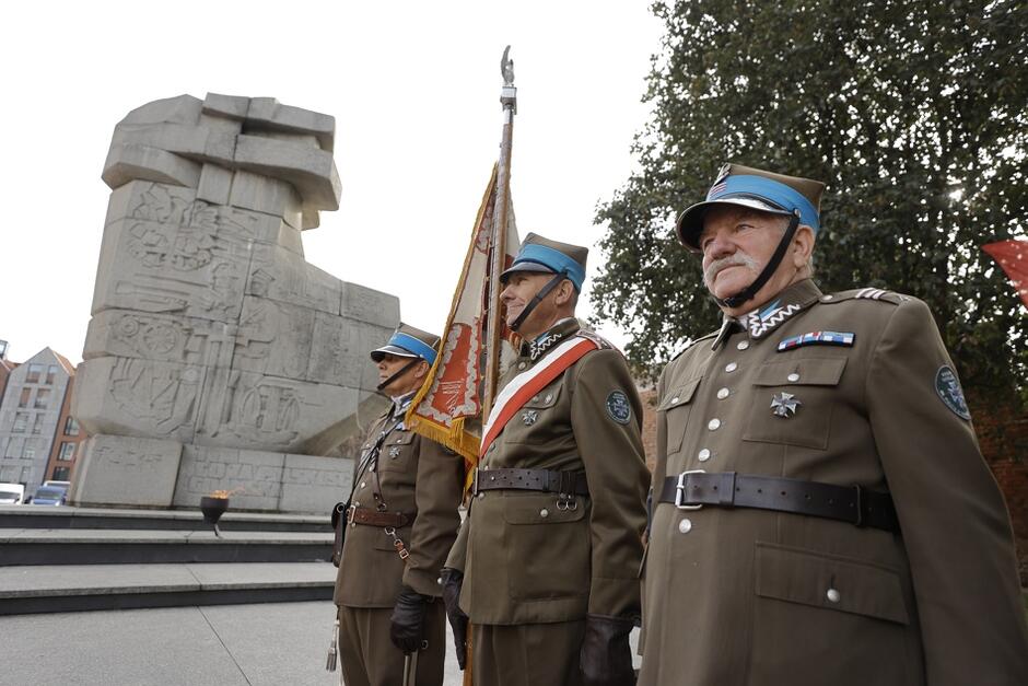 grupa mężczyzn w mundurach stoi w poczcie sztandarowym, w tle kamienny pomnik