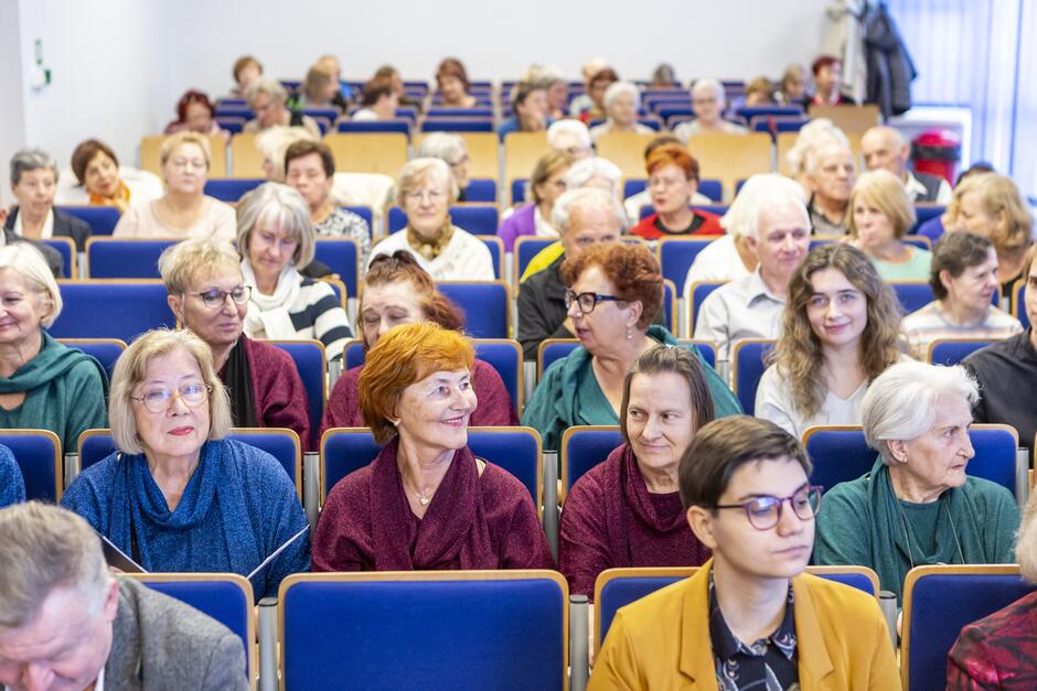 na zdjęciu kilkadziesiąt starszych osób, siedzi na krzesłach, w kilkunastu rzędach, patrzą przed siebie
