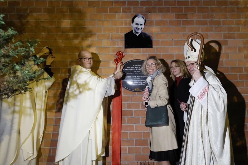 dwóch duchownych i dwie kobiety wieczorem, odsłaniają tablicę na murze