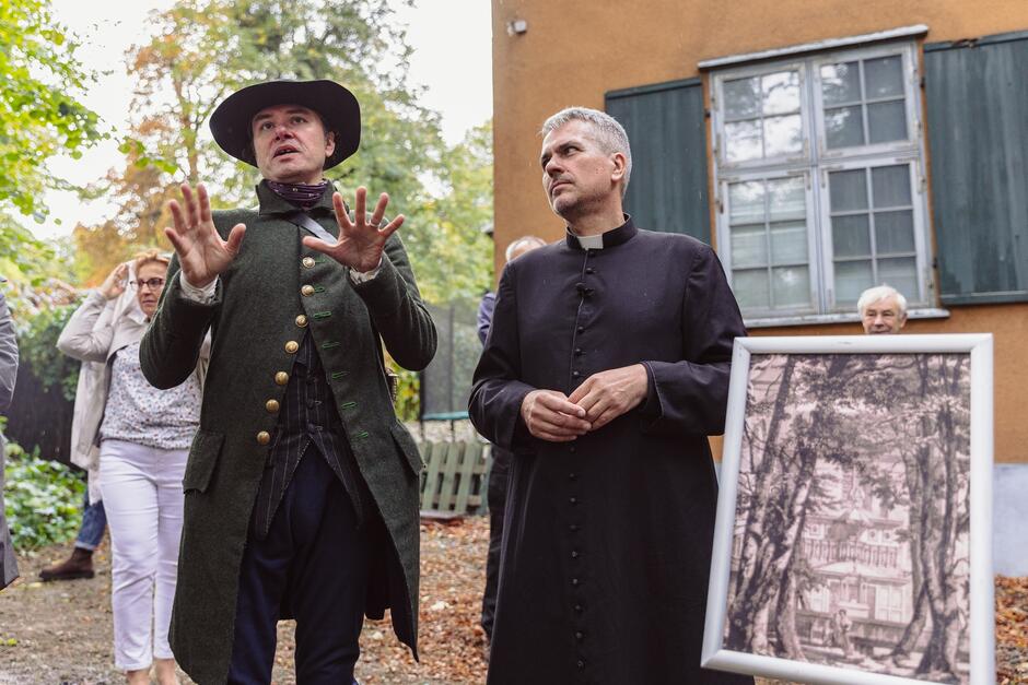 Dwaj mężczyźni przemawiają. Ten po lewej ubrany jest w strój mieszczański z XVIII wieku, po praweju stoi ksiądz w sutannie
