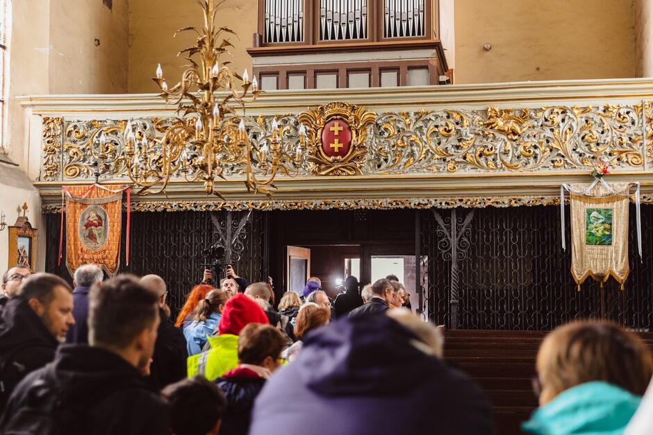 Wnętrze kościoła, widać głowy zwiedzających, ponad nimi - na chórze - widoczny jest kolorowy i ozdobny herb Gdańska