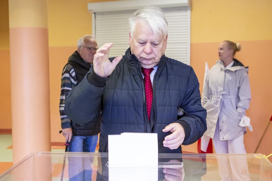 Mężczyzna po sześćdziesiątce, o siwych włosach, wrzuca kartę do urny wyborczej 
