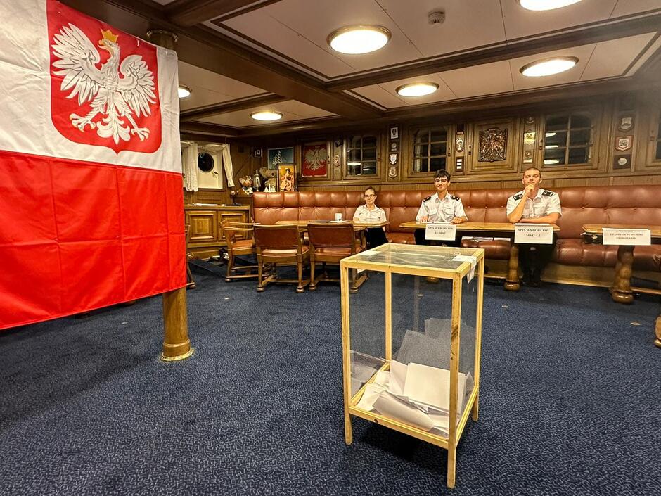 Po lewej bandera biało-czerwona, w środku urna wyborcza na drewnianych nogach, w tle trzy osoby siedzą za stołem 