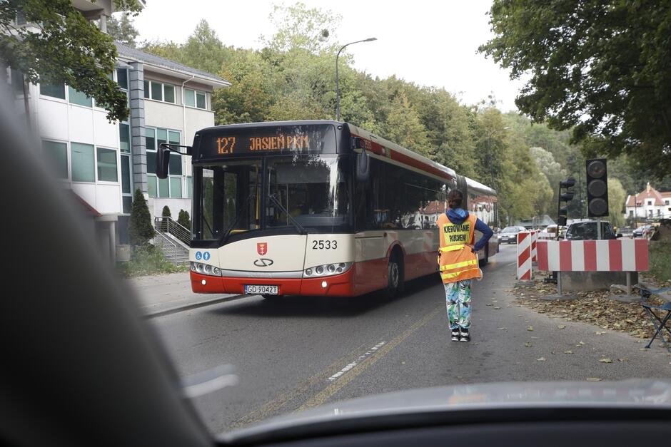 skrzyżowanie z ruchem sterowanym ręcznie, kobieta w kamizelce z napisem ruch wahadłowy stoi na ulicy, obok niej przejeżdża autobus